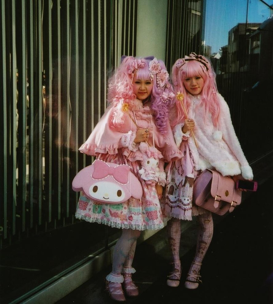 Two young women in pink Lolita fashion, posing outdoors with accessories. They wear elaborate costumes and carry matching handbags.