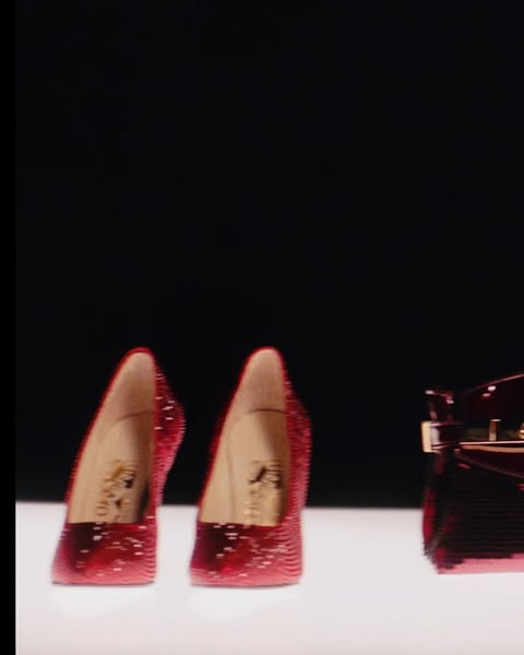 Two red, sparkling high heel shoes and a red patent leather handbag are positioned on a white surface against a black backdrop.