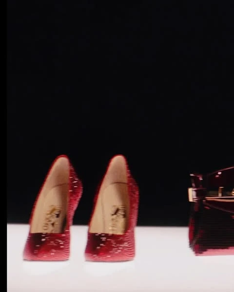 Two red, sparkling high heel shoes and a red patent leather handbag are positioned on a white surface against a black backdrop.