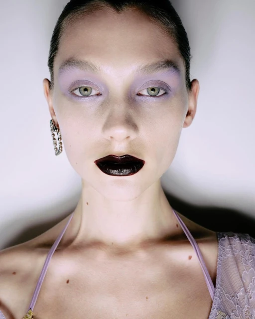 Close-up of a young woman with striking makeup, including dark lipstick and lavender eyeshadow, accentuated by dangling earrings.
