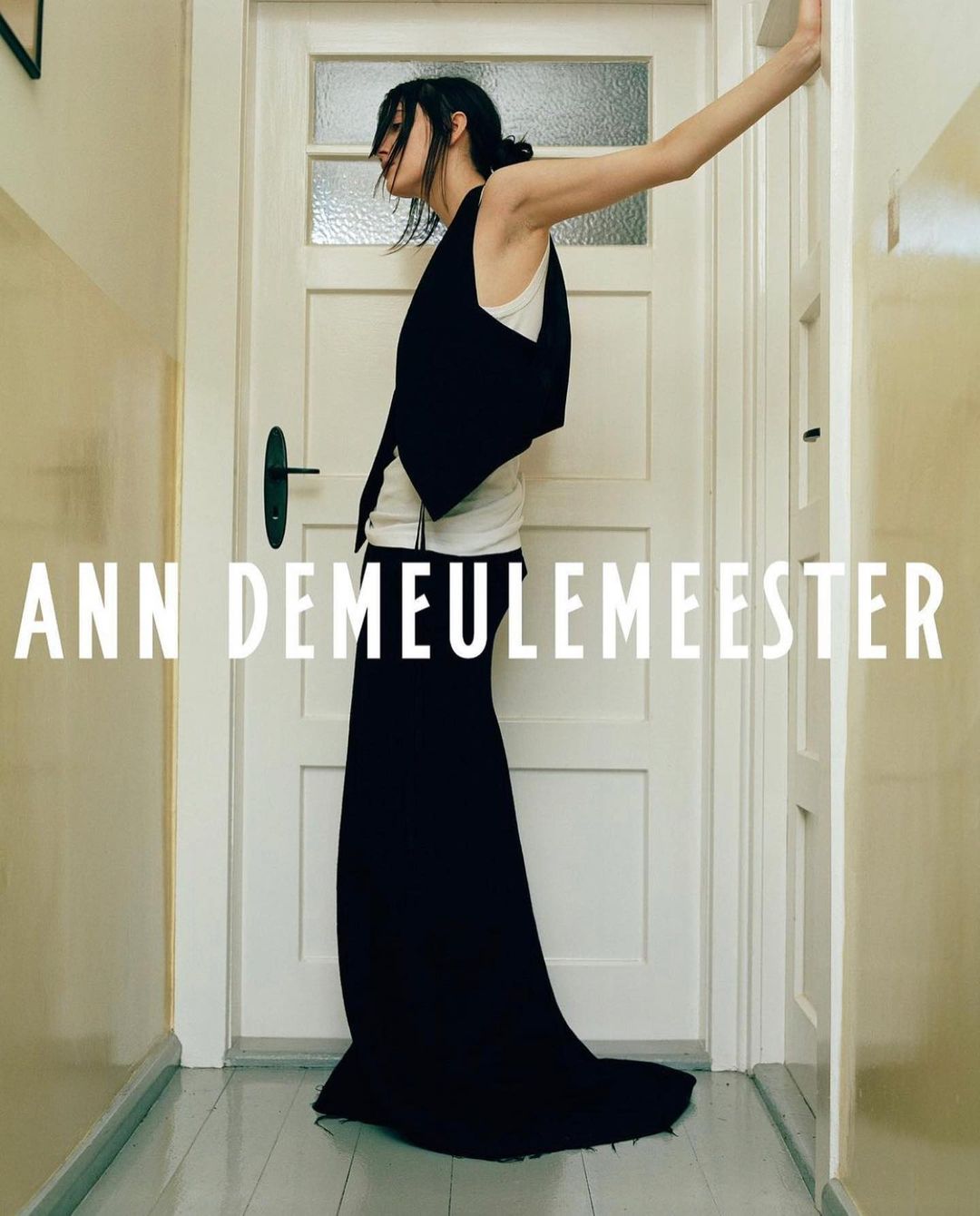 A woman stands in front of a white door wearing a black vest and long black skirt, with the words 'ANN DEMEULEMEESTER' written on the image.