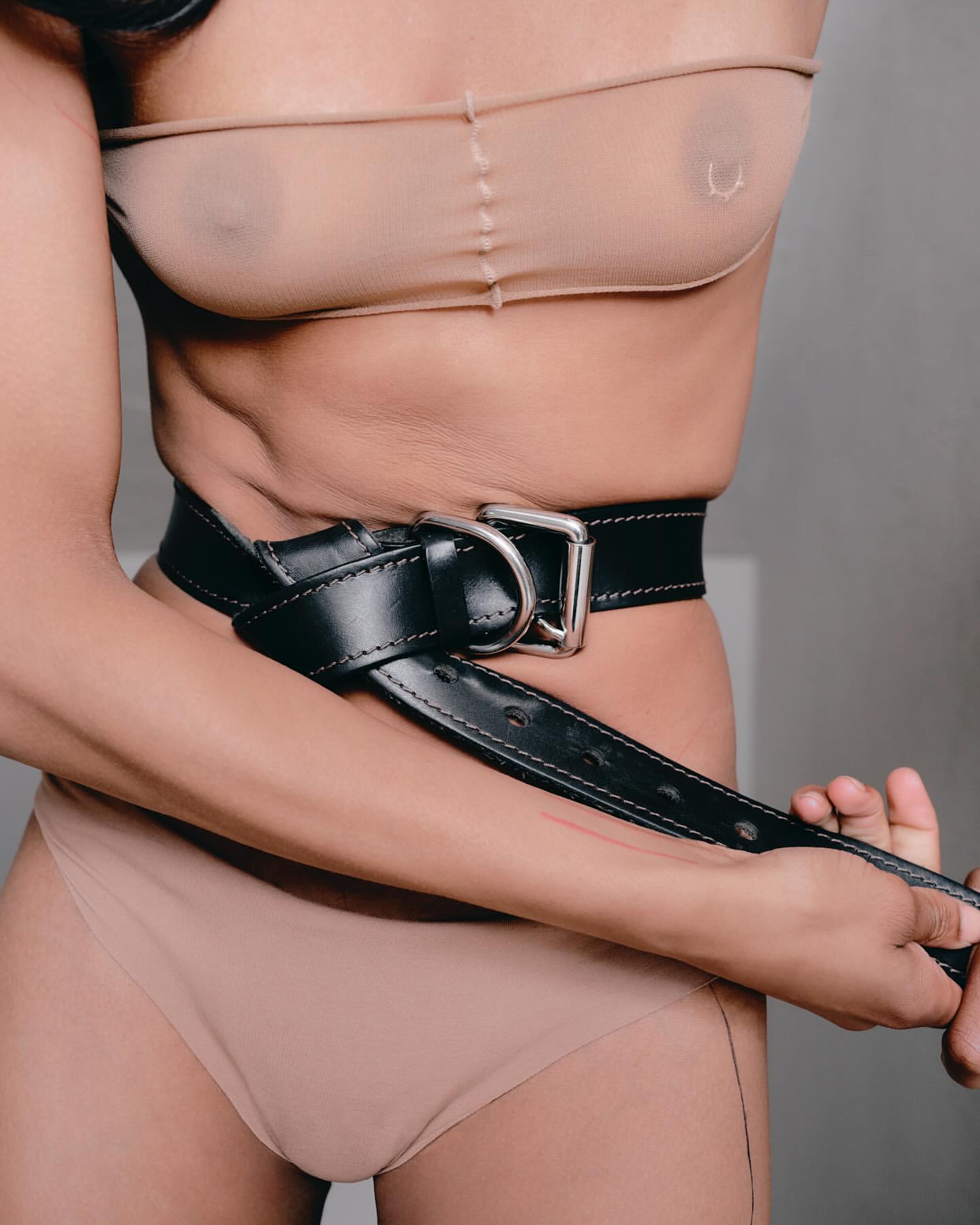 A woman is wearing a beige bra and underwear with a black leather belt around her midriff against a plain light gray background.