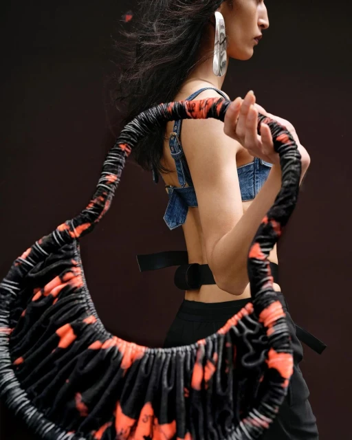 Runway fashion shot: a stylish adult female model presents a black and orange tie-dye handbag against a dark, neutral backdrop.