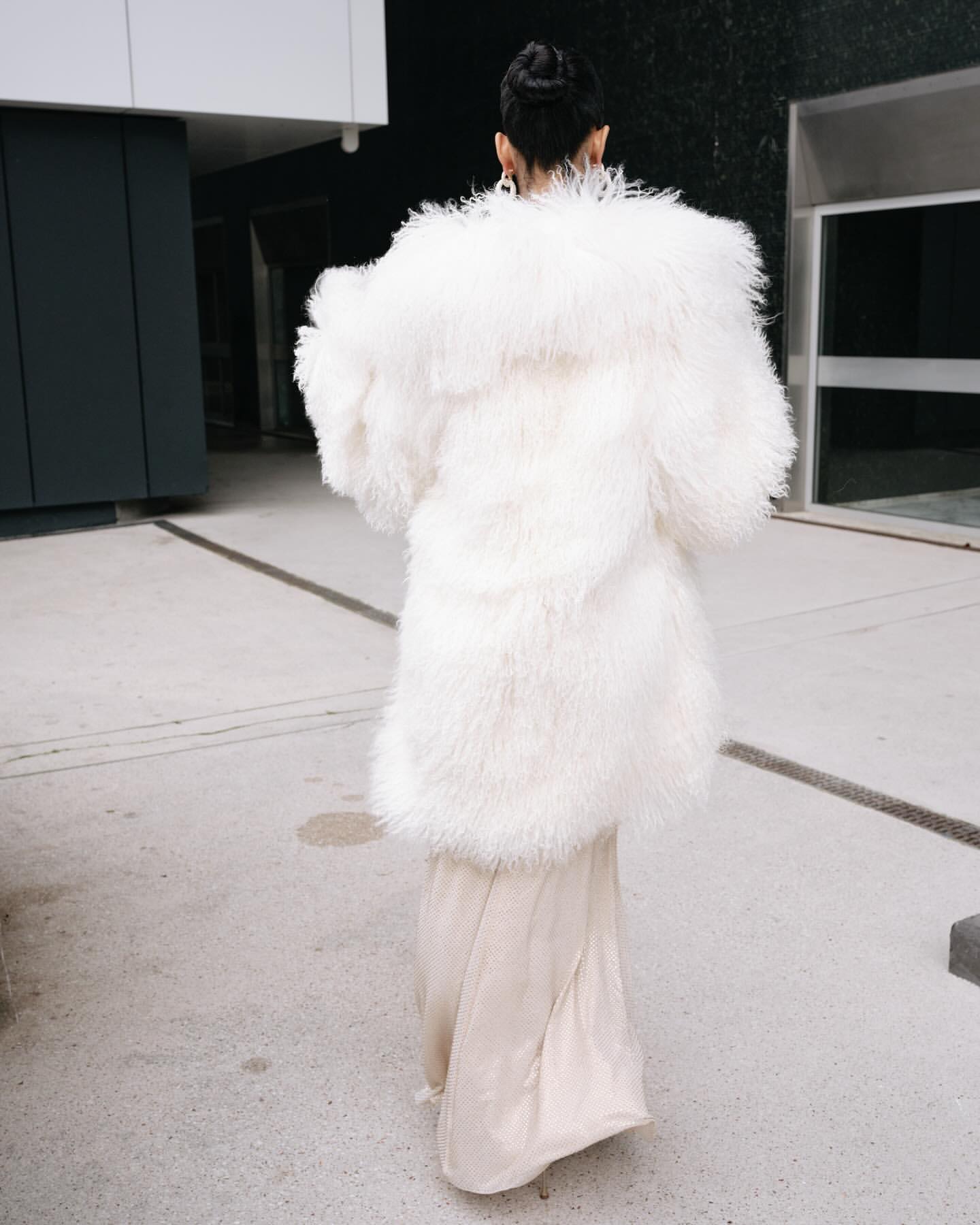 Woman in cream gown and oversized white fur coat walks on concrete. Back view shows fashion statement with texture contrast.
