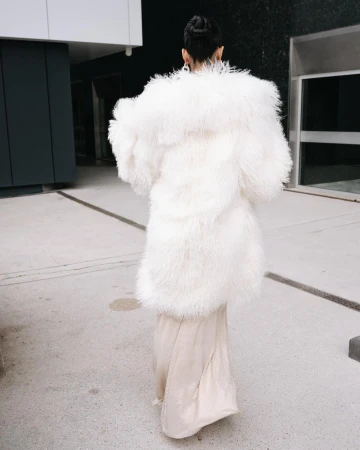 Woman in cream gown and oversized white fur coat walks on concrete. Back view shows fashion statement with texture contrast.