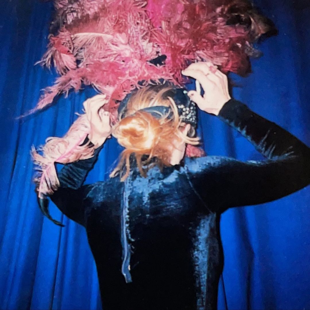 Woman in dark dress adjusts ornate feather headdress. Pink and red feathers frame her face against a blue draped curtain backdrop.