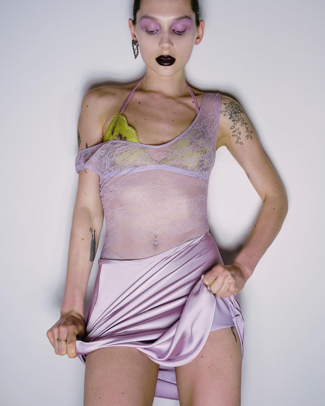 An adult female model poses in a photo studio wearing a lace top and lavender skirt, set against a neutral-toned background.