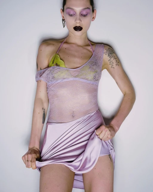 An adult female model poses in a photo studio wearing a lace top and lavender skirt, set against a neutral-toned background.