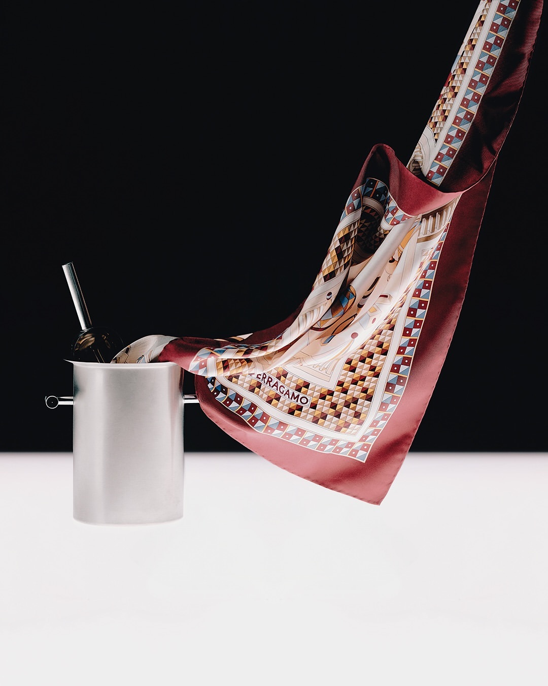 Elegant Ferragamo silk scarf draped artistically from silver ice bucket in a studio setting with black background.