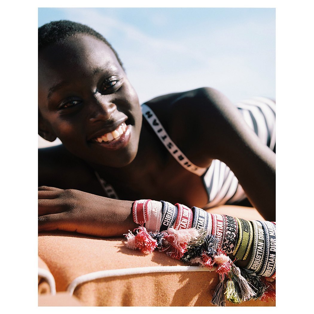 Close-up shot of a woman with dark hair smiling while lying on a beach towel, adorned with Christian Dior bracelets under a blue sky.