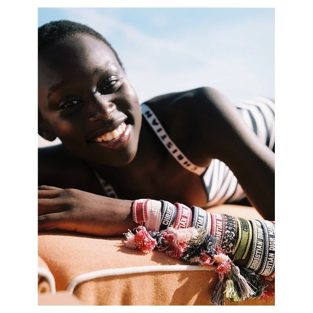 Close-up shot of a woman with dark hair smiling while lying on a beach towel, adorned with Christian Dior bracelets under a blue sky.