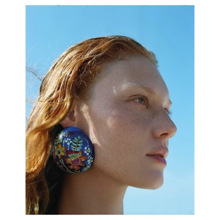 Close-up profile of a woman with red hair and freckles wearing a large round blue hand-painted earring against a clear sky.