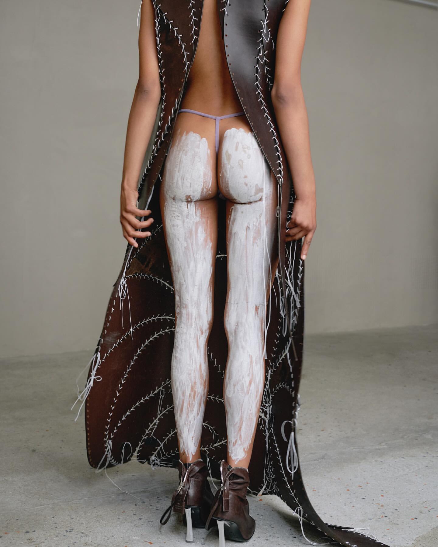 Model seen from behind wearing an open-back brown leather garment with white stitching, with white paint on legs.