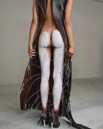 Model seen from behind wearing an open-back brown leather garment with white stitching, with white paint on legs.