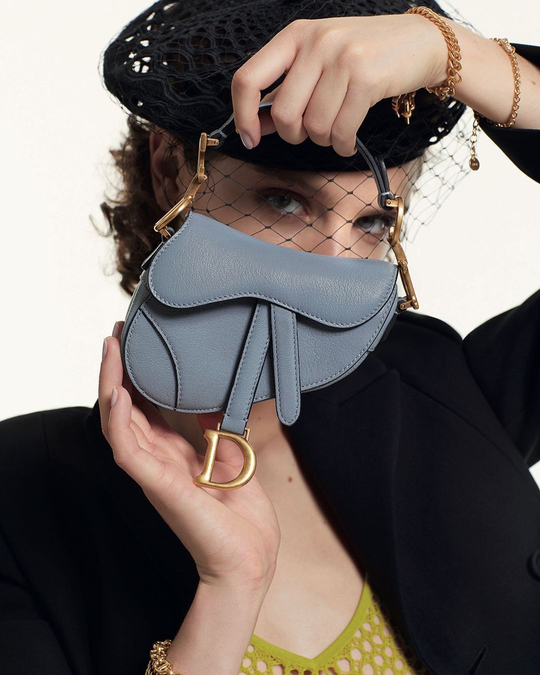 An adult woman poses in a photo studio with a light blue Dior purse in her hands, wearing a black net hat and a black jacket.