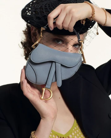 Woman wearing a black netted beret and jacket holding a light blue leather saddle handbag up to her face, photographed by Alice Rosati.