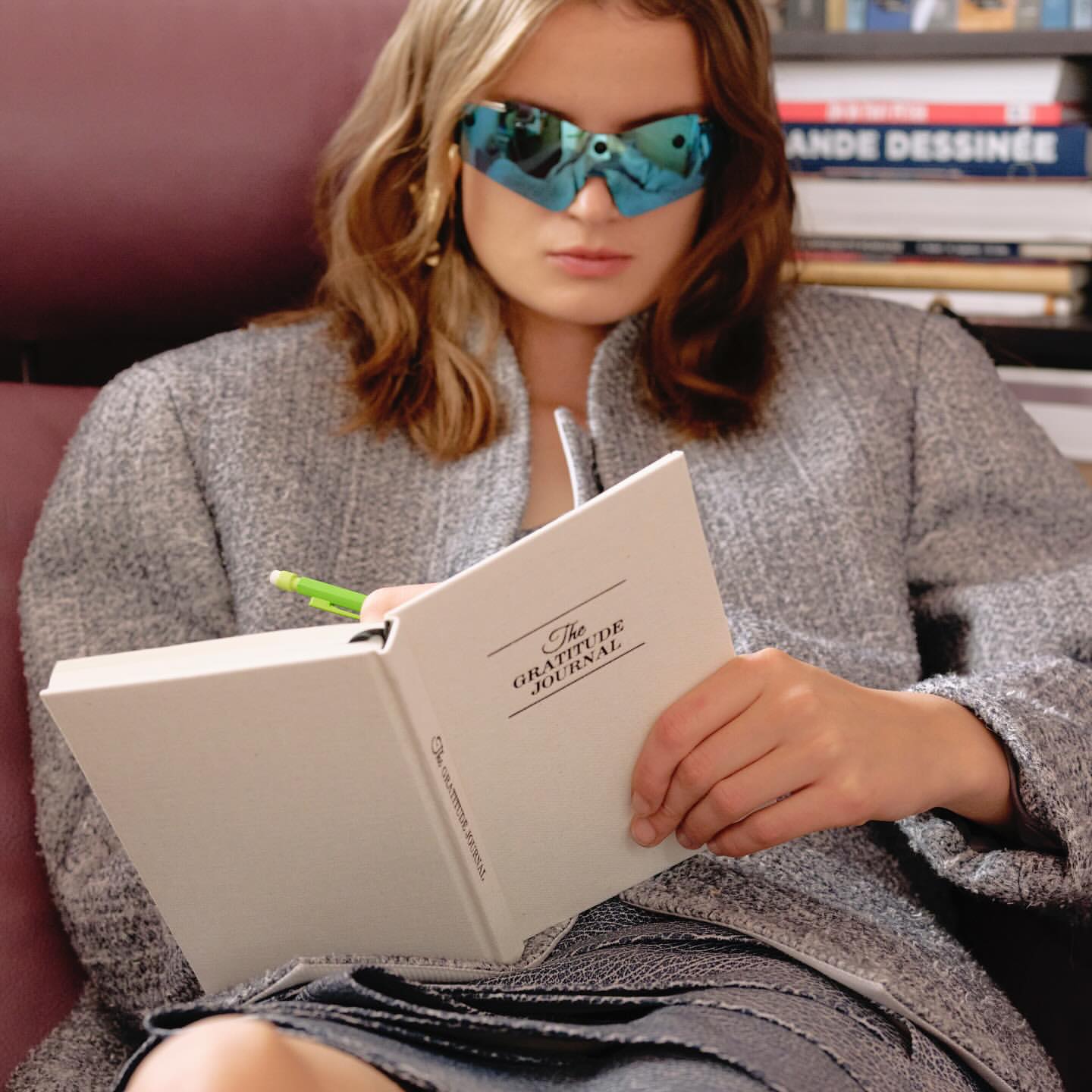Woman in sunglasses writing in journal in indoor setting with books in background, captured in a medium shot with soft lighting.
