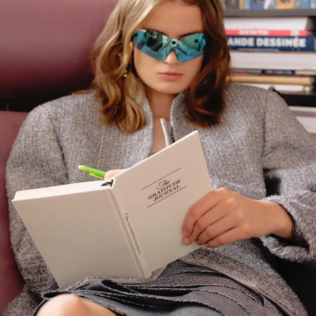 Woman in sunglasses writing in journal in indoor setting with books in background, captured in a medium shot with soft lighting.