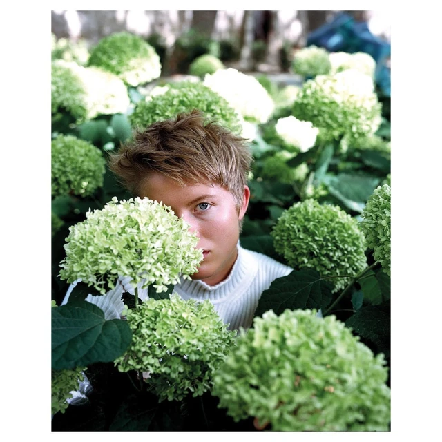 Portrait of a young adult with blond hair among green hydrangeas. Person wearing a ribbed turtleneck, posing outdoors in natural light.