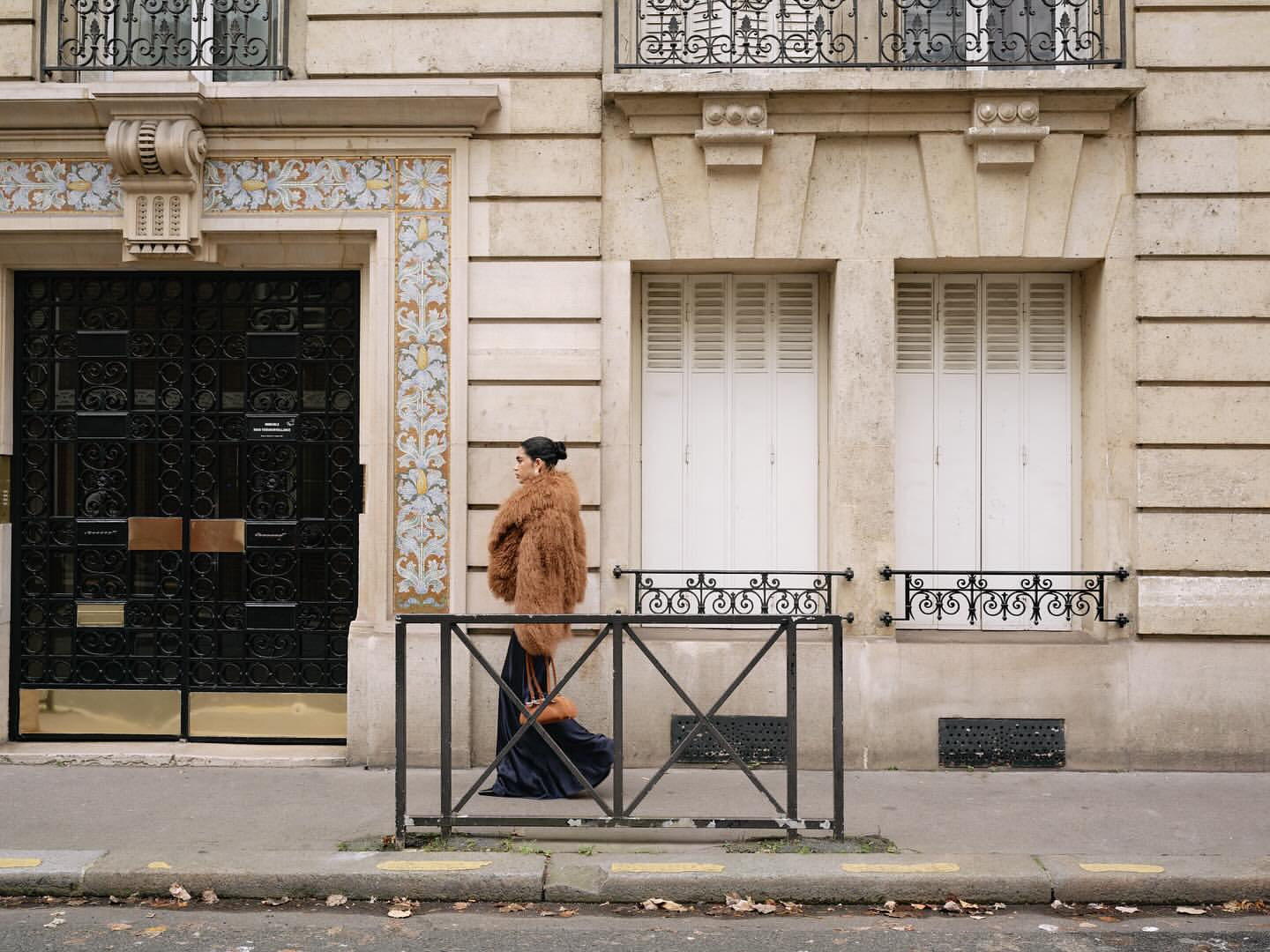Woman in fur coat walks on city street in front of ornate building. Fashion editorial photography with neutral tones and European architecture.