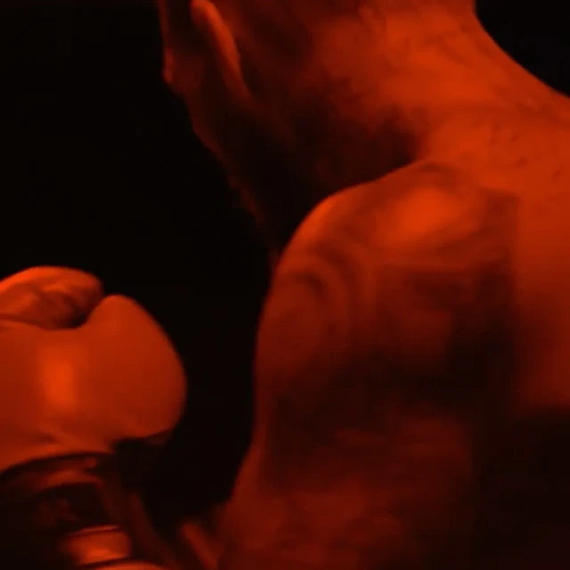Close-up of a male boxer's torso and back showing muscular physique and tattoo details, with boxing glove visible in low red lighting.