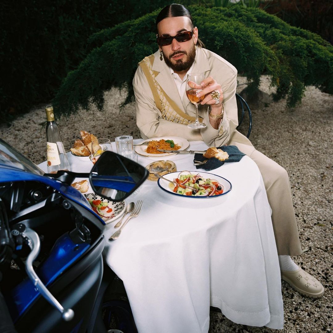 Man with beard and sunglasses seated at a table with food, holding a glass of wine, beside a blue motorcycle