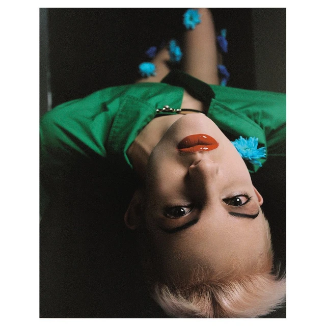 Close-up portrait of a fair-skinned person with short blond hair, red lipstick, and a green top, lying down with blue flowers scattered around.