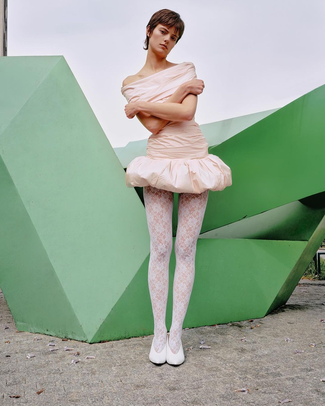 Young woman in an off-the-shoulder pastel dress stands outdoors in white tights and ballet flats next to a green geometric structure.
