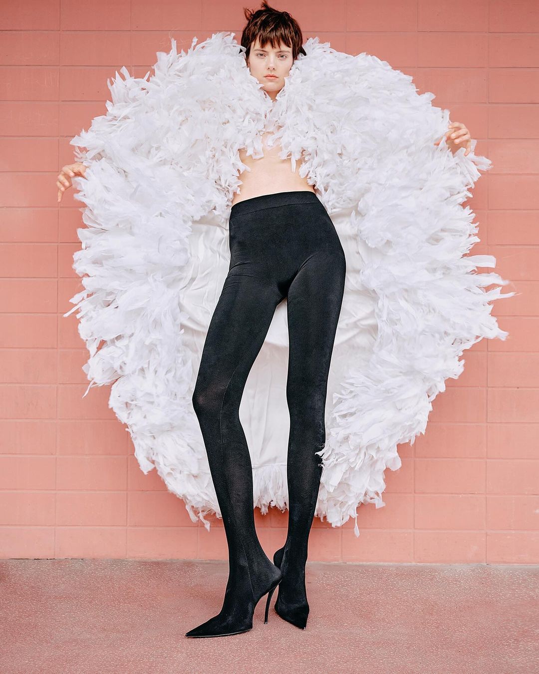 Fashion model in black leggings and stiletto heels poses in front of a pink wall wearing a large white feather boa.