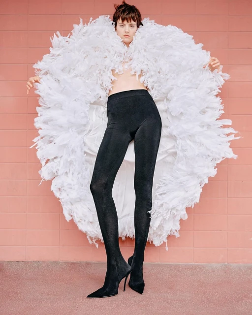 Fashion model in black leggings and stiletto heels poses in front of a pink wall wearing a large white feather boa.