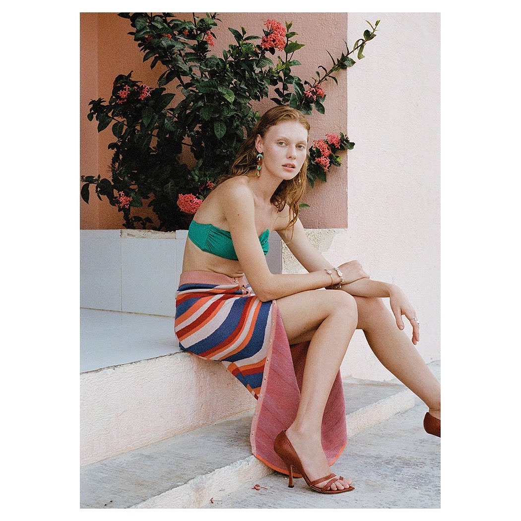 Woman in swimwear and striped skirt sitting on steps outdoors with flowering bush. Wearing jewelry, high heel shoes, looking at camera.