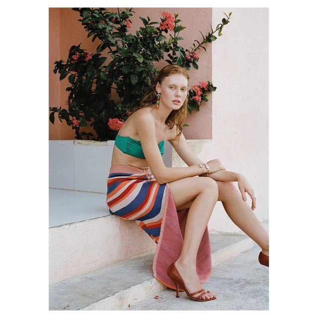 Woman in swimwear and striped skirt sitting on steps outdoors with flowering bush. Wearing jewelry, high heel shoes, looking at camera.