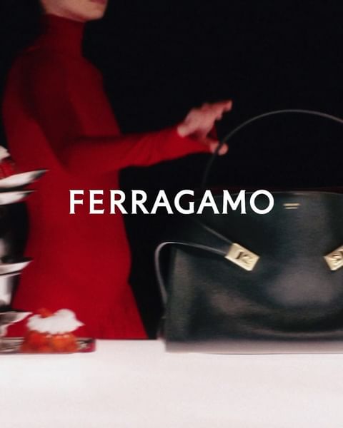 Ferragamo handbag shown with a figure in a red turtleneck jumpsuit and artful fruit display on a white table against a black backdrop.