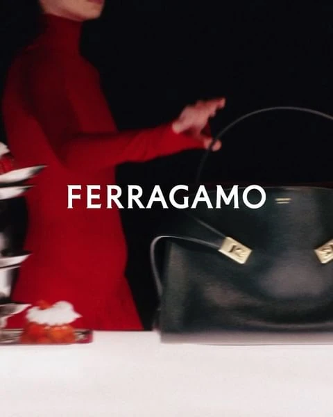 Ferragamo handbag shown with a figure in a red turtleneck jumpsuit and artful fruit display on a white table against a black backdrop.