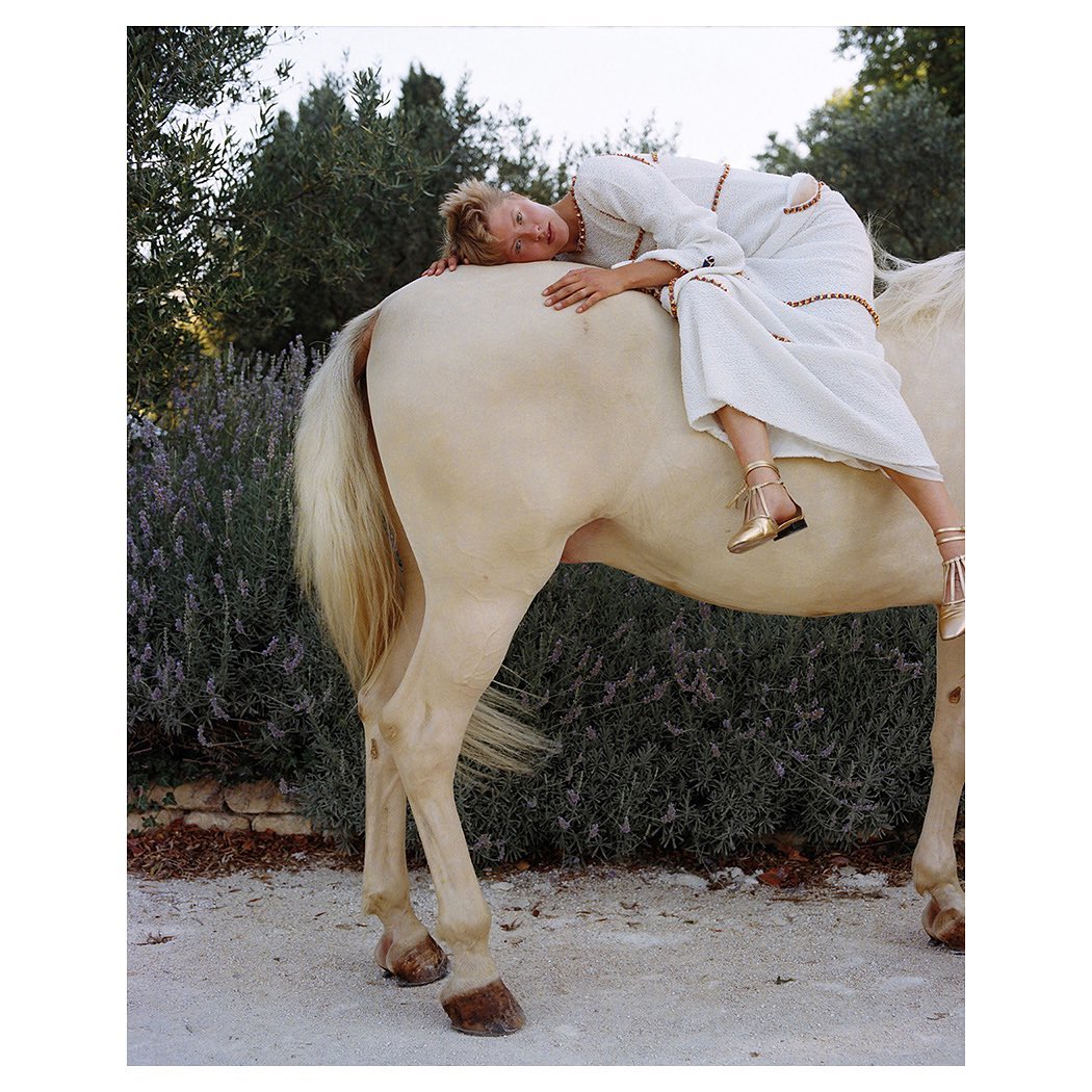 Woman in white dress lying on the back of a horse in an outdoor setting with green trees and plants in the background.
