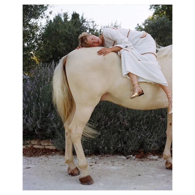 Woman in white dress lying on the back of a horse in an outdoor setting with green trees and plants in the background.