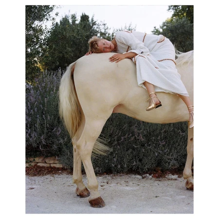 Woman in white dress lying on the back of a horse in an outdoor setting with green trees and plants in the background.