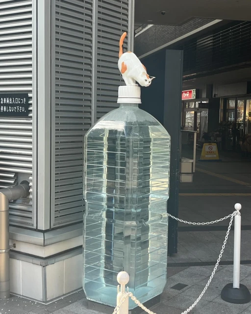 A playful cat sculpture perches on a large, clear water bottle against an urban backdrop with an entranceway and metal siding.