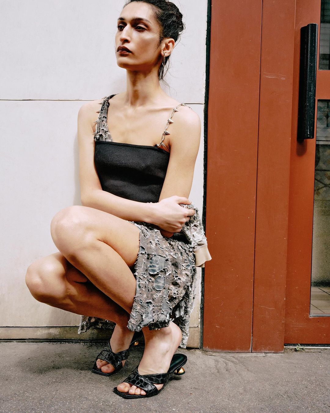 A woman in a black top and patterned skirt poses in an urban setting. She is crouching with her arms crossed and wearing heeled sandals.