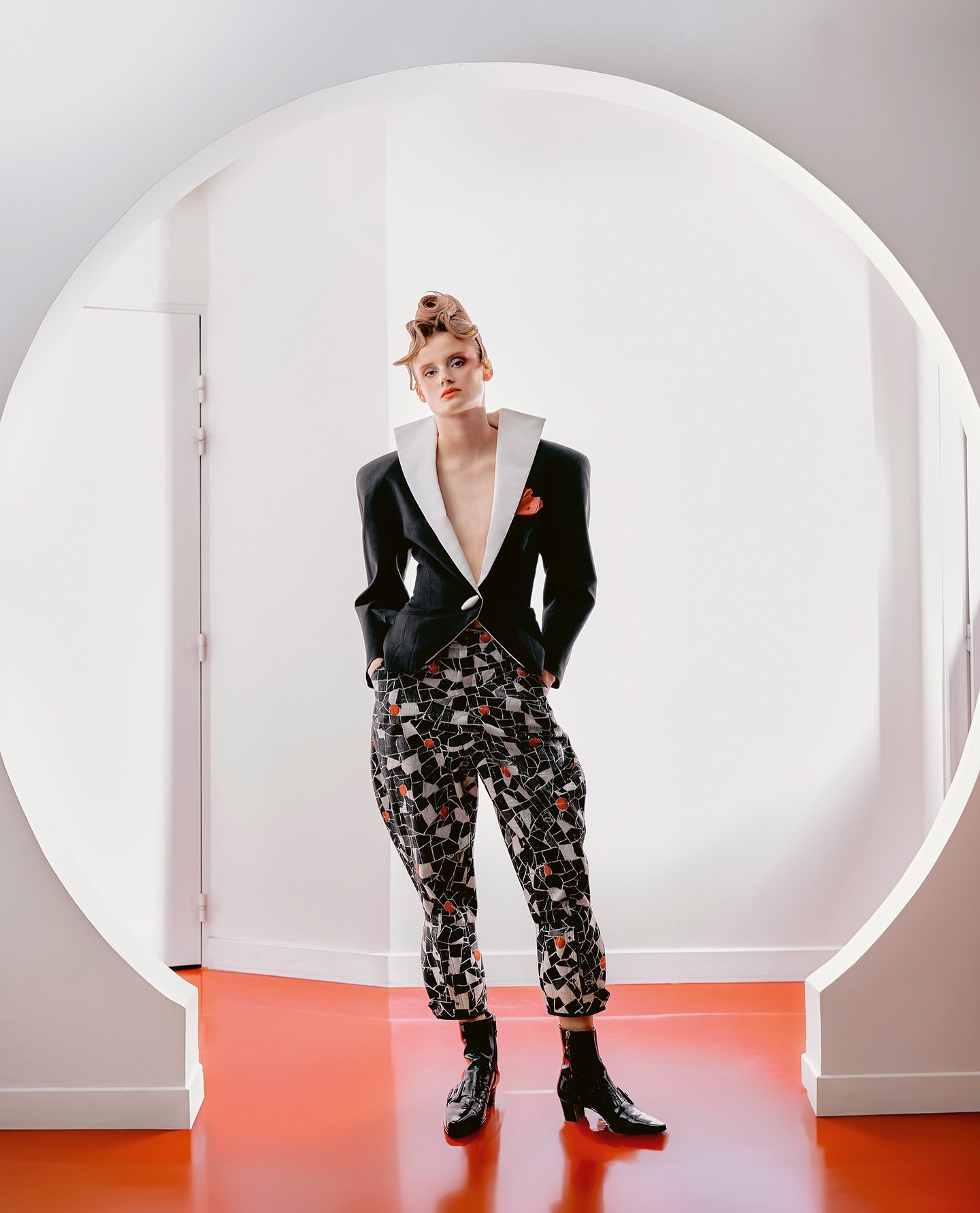 Fashionable woman in a black blazer with statement collar and geometric patterned pants, posing indoors on a red floor against a white wall.