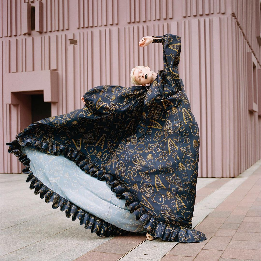 Woman in oversized patterned dress poses in front of a pink building. The long dress flows around her as she looks up with her arm raised.