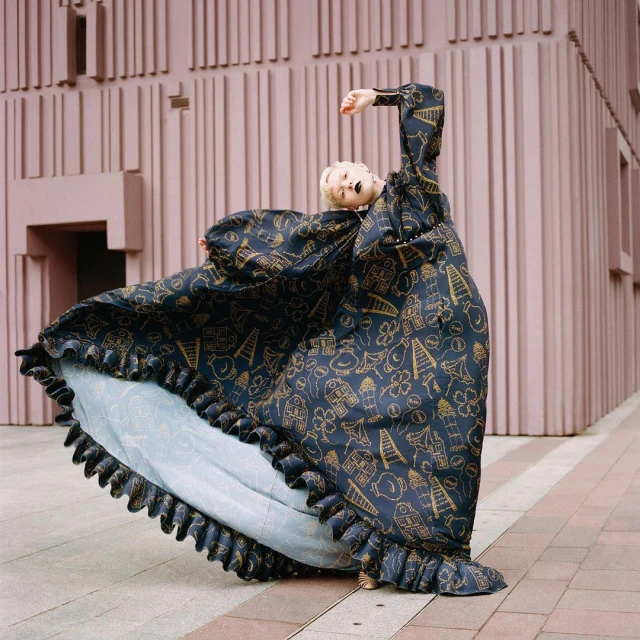 Woman in oversized patterned dress poses in front of a pink building. The long dress flows around her as she looks up with her arm raised.