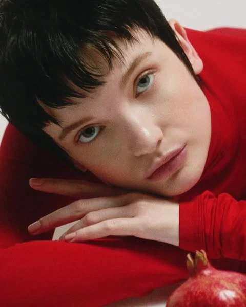 Close-up portrait of a young adult with short dark hair and light eyes, wearing a red turtleneck, with a pomegranate in the foreground.