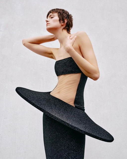 A woman in a glittering black two-piece dress with a large pointed element extending from the hip, posing against a light grey wall.