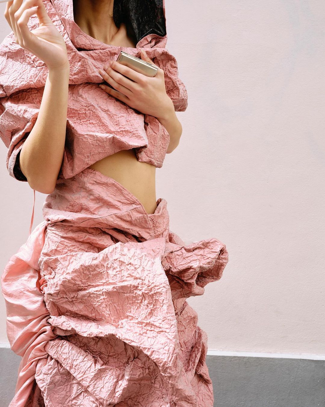 Woman poses in a rose-colored, textured garment. The material has a crinkled effect, resembling crushed paper. High fashion studio shot.