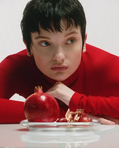 Close-up portrait of a woman with short black hair in a red top, alongside a pomegranate and grapes on a clear dish.