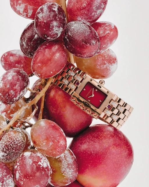 Close-up of a rose gold wristwatch with a red face, set against a background of red grapes dusted with white powder. 