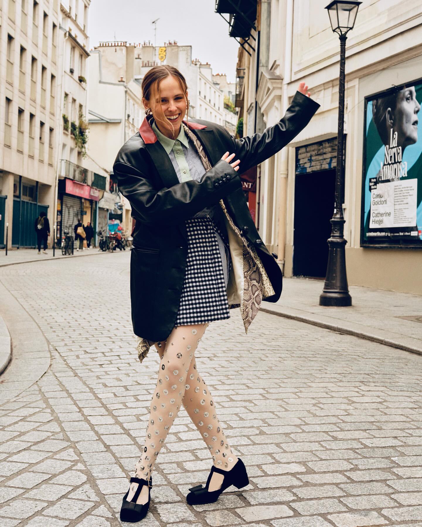 Smiling woman in a black leather jacket and patterned skirt dancing on a cobblestone Paris street by photographer Alice Rosati