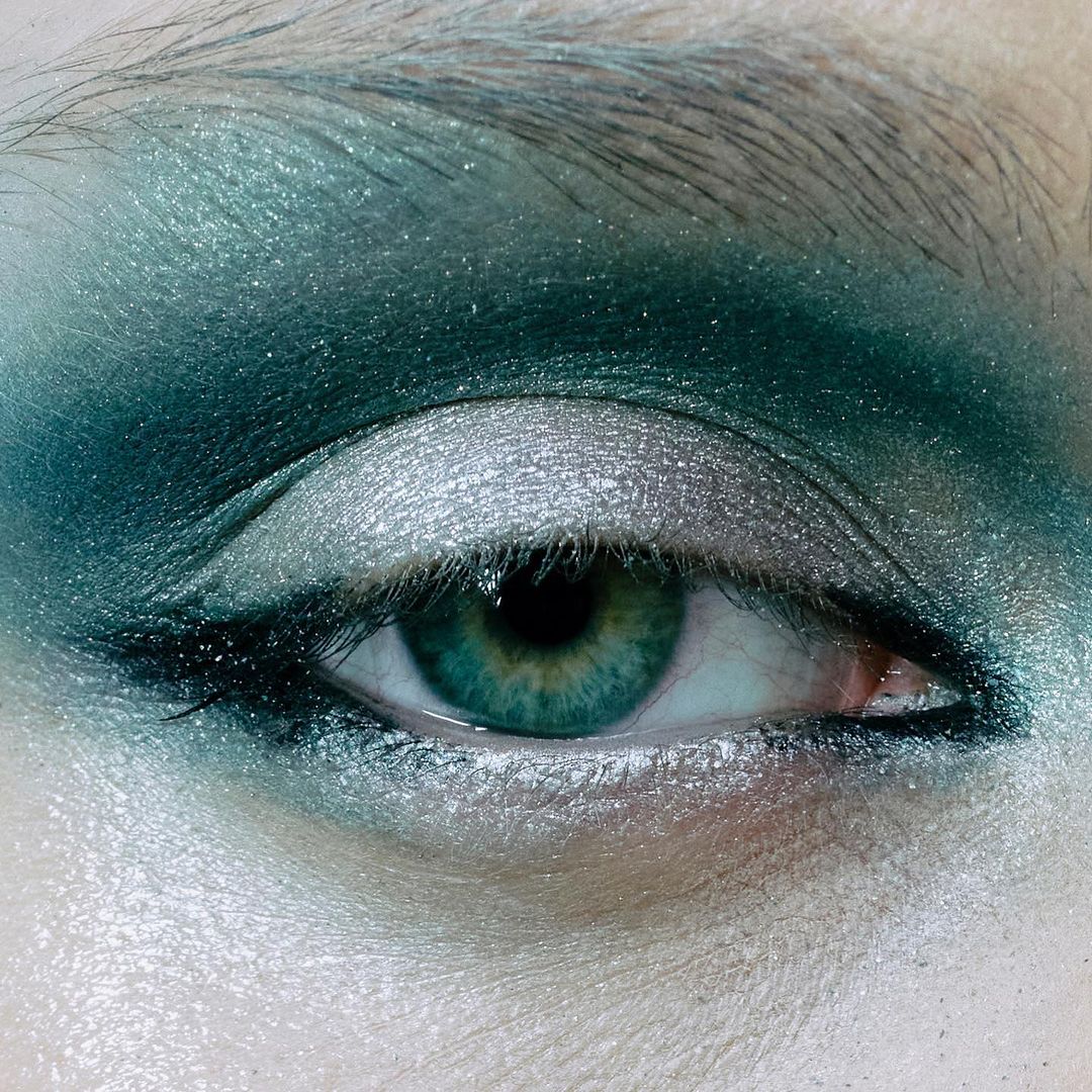 Close-up of a woman's eye featuring vivid green irises and expertly applied emerald and silver eyeshadow for a striking beauty look.