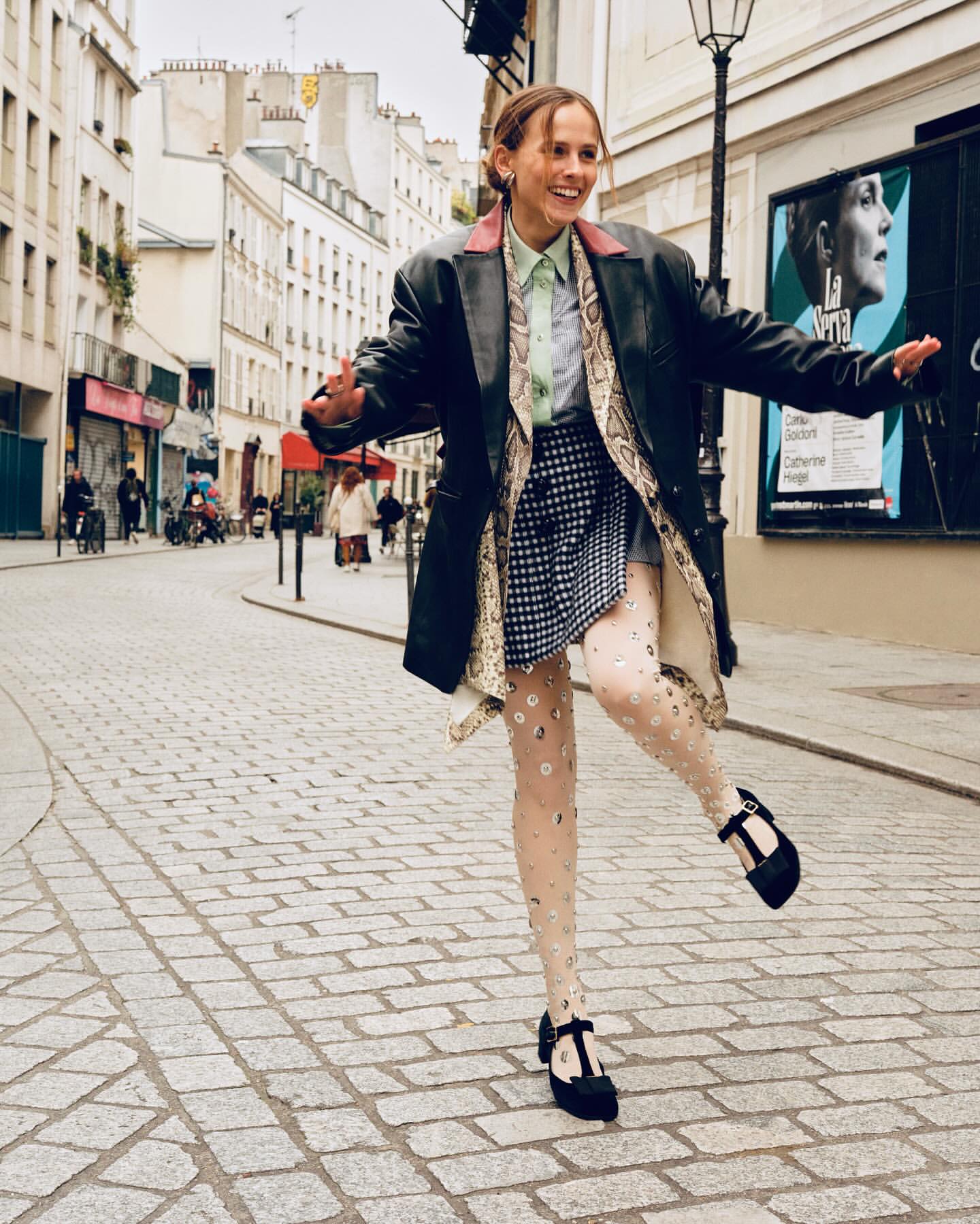 A smiling young woman in a black leather jacket poses on a cobblestone street in Paris, France, wearing patterned tights and Mary Jane shoes.